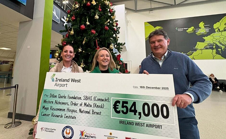 Three people stand in front of a Christmas tree, smiling whilst holding a large cheque for €54,000 from Ireland West Airport, dated 16 December 2025. The donation supports several charities, including Mayo Roscommon Hospice.