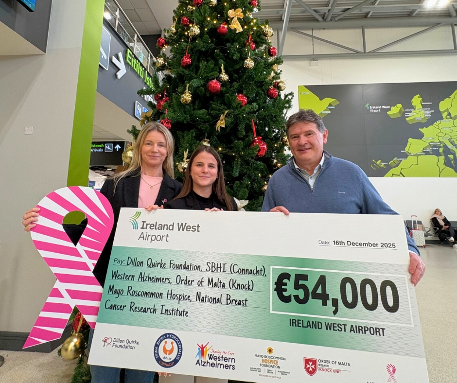 Three people stand by a Christmas tree in an airport, holding a large cheque for €54,000 from Ireland West Airport, dated 16 December 2025, benefiting various charities including Mayo Roscommon Hospice.