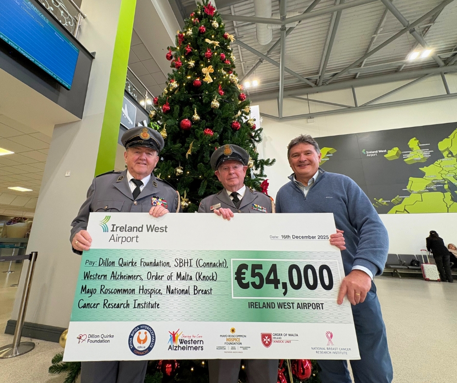 Three men stand in front of a Christmas tree, holding a large €54,000 charity cheque from Ireland West Airport, with logos of several foundations and organisations—including Mayo Roscommon Hospice—visible.