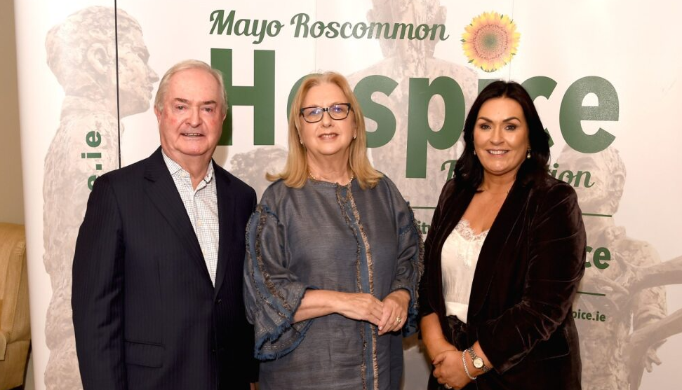 Three adults stand in front of a Mayo Roscommon Hospice Foundation banner, smiling at the camera. Representing the Mayo Roscommon Hospice community, two women in formal attire and one man in a suit appear to be attending an indoor event or gathering.