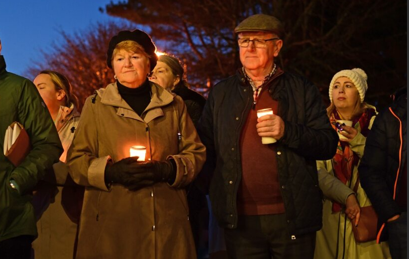 A group of people stand outside at dusk, holding candles during a vigil for the Mayo Roscommon Hospice. Most are dressed warmly in coats and hats, and they appear solemn and reflective. Trees are visible in the background.
