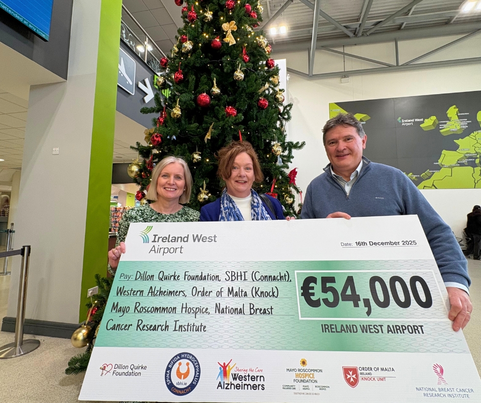 Three people stand in front of a decorated Christmas tree, smiling and holding an oversized cheque for €54,000 from Ireland West Airport, made out to various charities, including Mayo Roscommon Hospice. The donation date is 16 December 2025.