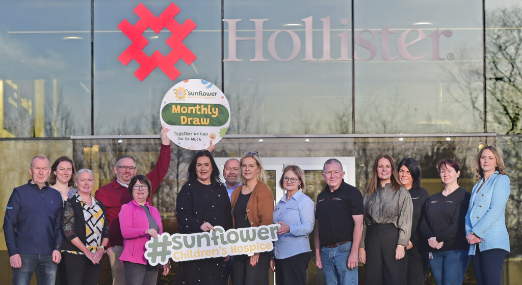 A group of people stand outside a Hollister building, holding signs for the Sunflower Children’s Hospice Monthly Draw, which supports causes like the Mayo Roscommon Hospice. Some are smiling, and the group is gathered in front of large windows.