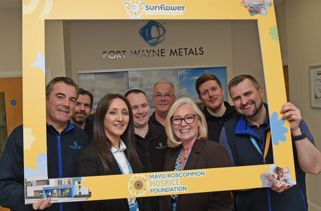 Eight people smile inside a building, holding a large yellow photo frame labelled Mayo Roscommon Hospice Foundation with a sunflower logo and Sunflower Children’s Hospice at the top. A Fort Wayne Metals sign is visible behind them, highlighting their support for the Mayo Roscommon Hospice’s mission.
