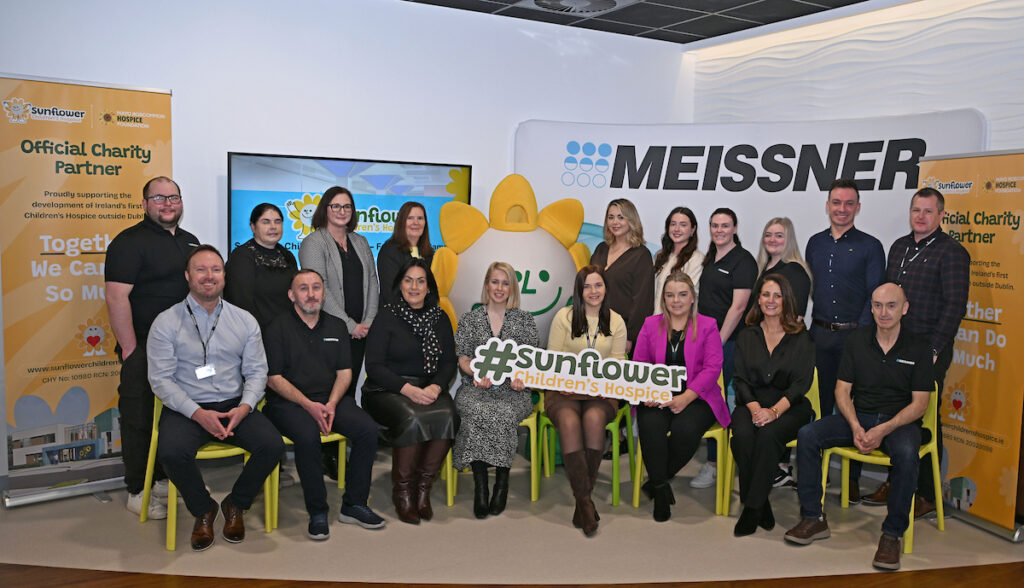 A group of people pose indoors with Sunflower Children’s Hospice banners, a sunflower mascot, and a Meissner sign. Smiling and sitting or standing in two rows, they are supporting a charity partnership inspired by the values shared with Mayo Roscommon Hospice.