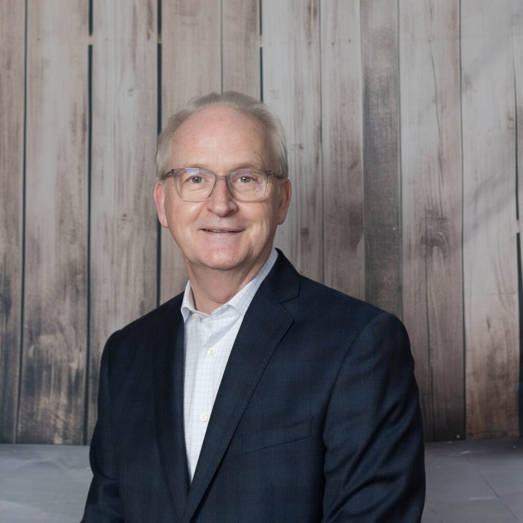 A man with short grey hair and glasses, wearing a dark suit jacket and light shirt, stands in front of a wooden panel background, smiling at the camera—possibly attending an event in support of Mayo Roscommon Hospice.