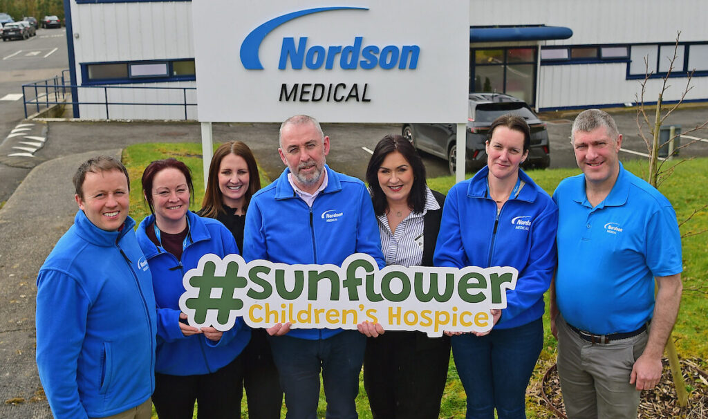 Seven people in blue jackets stand outside a Nordson Medical building, smiling and holding a sign that reads #sunflower Children's Hospice, showing their support for both the Mayo Roscommon Hospice and local children’s services.