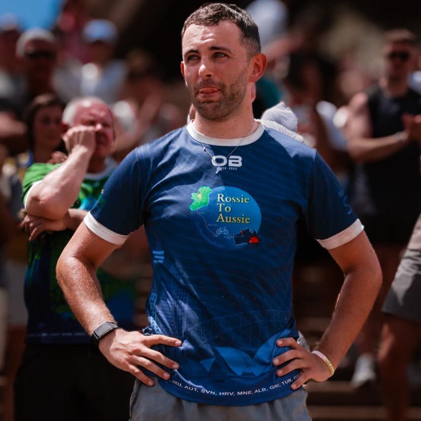 A man in a blue Rossie To Aussie shirt stands with hands on hips, looking ahead, perhaps at a Mayo Roscommon Hospice event. People applaud in the blurred background on stone steps.