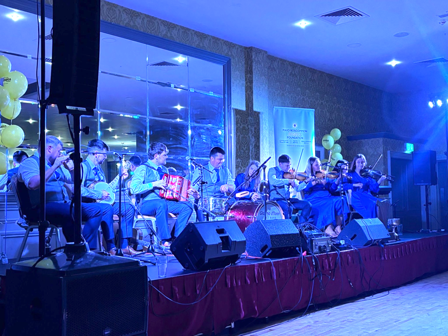 A band of musicians playing traditional instruments, including fiddles, banjo, and accordion, perform on a stage in a blue-lit room with yellow balloons and a mirrored wall in the background, as part of a fundraising event for Mayo Roscommon Hospice.