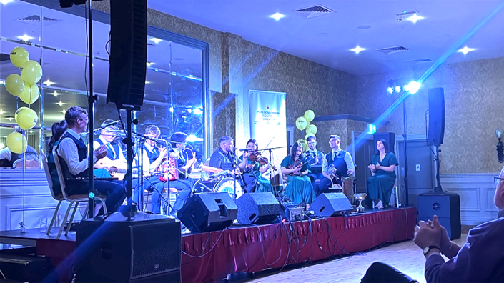 A band plays traditional instruments on a stage adorned with yellow balloons and blue lighting, whilst people watch from the audience in support of Mayo Roscommon Hospice. Some musicians wear hats and waistcoats, and there are microphones and speakers on stage.