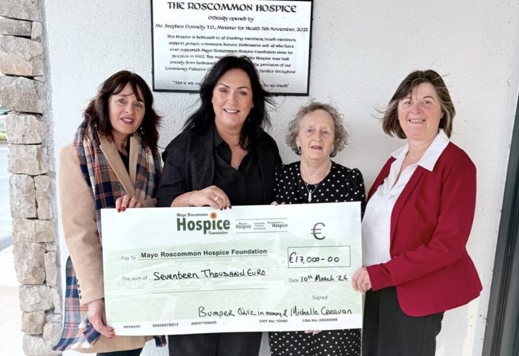 Four women stand in front of The Roscommon Hospice, smiling and holding an oversized cheque for €17,000 made out to Mayo Roscommon Hospice Foundation. A commemorative plaque is visible on the wall behind them, highlighting their support for the Mayo Roscommon Hospice.