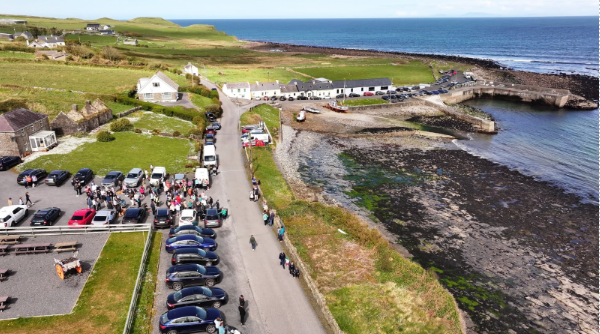 Aerial view of a coastal village with parked cars, people gathering near buildings—some perhaps attending a Mayo Roscommon Hospice event—green fields, rocky shoreline, and the sea in the background under a partly cloudy sky.