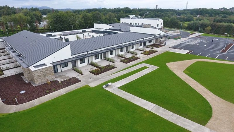 Aerial view of a modern, single-storey building with a grey roof, surrounded by green lawns, paths, landscaped areas, and a large car park—this impressive facility is the Mayo Roscommon Hospice.