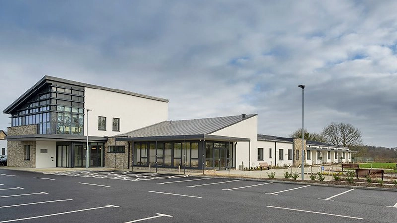 Modern, single-storey Mayo Roscommon Hospice building with large windows and a sloping roof, surrounded by a car park with marked spaces and some greenery. The sky is mostly cloudy.
