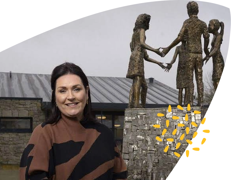 A woman stands smiling in front of the modern stone Mayo Roscommon Hospice building and a bronze statue of four people holding hands. Yellow leaf-like graphics are overlaid near the statue. The sky is overcast.