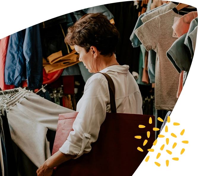 A woman with short brown hair, wearing a white shirt and carrying a large brown shoulder bag, shops for clothes in a shop filled with hanging garments, possibly browsing at a Mayo Roscommon Hospice charity shop.