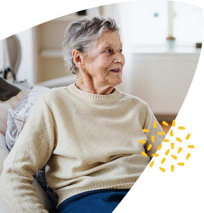 An older woman with grey hair, wearing a cream jumper, sits on a sofa and smiles. There is a small yellow decorative graphic near her side. The room is bright with soft natural light, creating a warm and comforting atmosphere reminiscent of the supportive care provided by Mayo Roscommon Hospice.