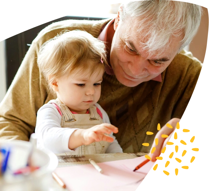 An older adult and a young child sit together at a table at the Mayo Roscommon Hospice, drawing or writing on pink paper with crayons. The adult smiles whilst watching the child, who is focused on her artwork.