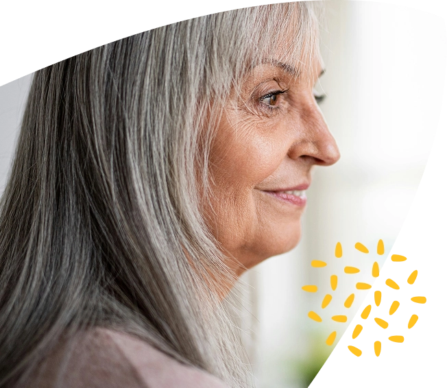 Older woman with long grey hair smiling in profile, with a neutral background and a yellow abstract design near the bottom right corner, representing the compassionate care offered by Mayo Roscommon Hospice.
