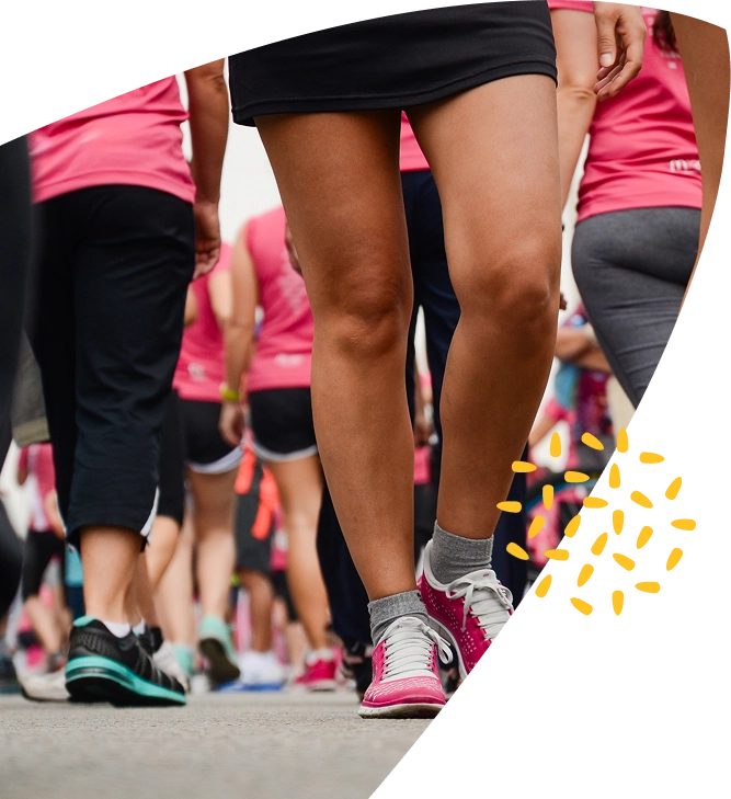 A group of people wearing sports clothes, mostly in pink tops, walk outdoors in support of Mayo Roscommon Hospice. The focus is on their legs and trainers as they move together. Yellow graphic dots decorate the lower right corner.
