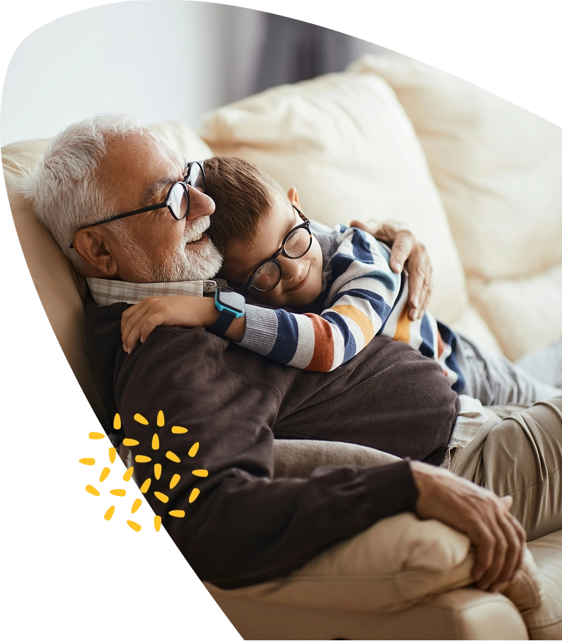 An elderly man and a young boy, both wearing glasses, sit on a beige sofa. The boy hugs the man warmly, and they both smile, appearing content and relaxed together—capturing a tender moment supported by the caring environment of Mayo Roscommon Hospice.