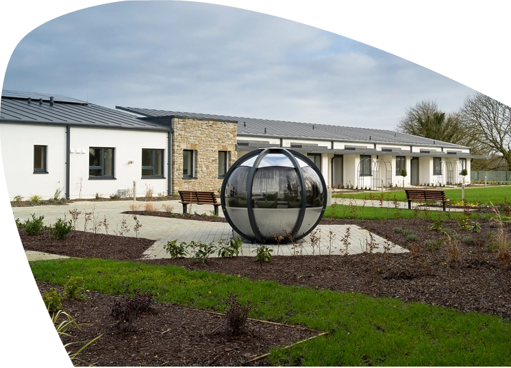 A modern single-storey building with a stone accent wall, surrounded by gardens and benches, serves as the Mayo Roscommon Hospice. A large metallic sphere sculpture sits in the centre of a paved area in front, creating a serene and welcoming atmosphere.