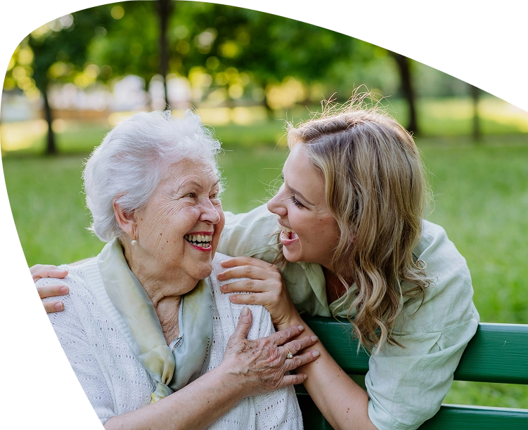 An elderly woman and a younger woman sit on a bench outdoors, smiling and laughing together, surrounded by green trees and grass, enjoying a peaceful afternoon supported by the Mayo Roscommon Hospice.
