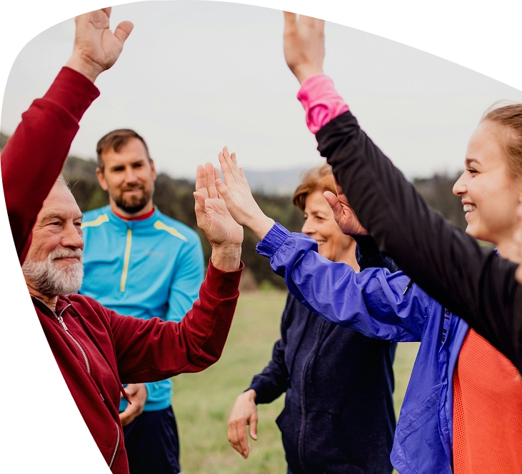 Four people outdoors, smiling and exchanging high fives. They are dressed in sporty, colourful jackets and appear to be in a park or open grassy area, with trees and hills visible in the background—capturing the spirit of community often seen at Mayo Roscommon Hospice events.