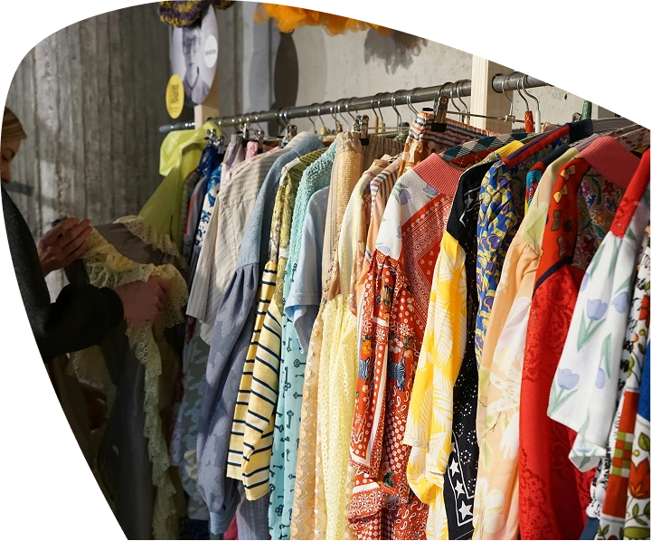 A person browses a clothing rail filled with colourful dresses and shirts in various patterns and styles, displayed against a concrete wall at the Mayo Roscommon Hospice charity shop.
