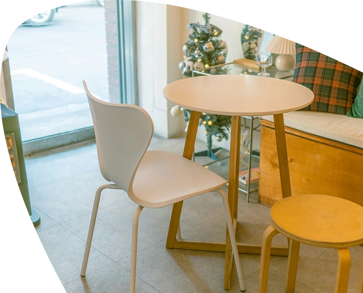 A white round table with a white chair and a small wooden stool beside it, near a window. A decorated Christmas tree and a cushioned bench are in the background, creating a warm atmosphere reminiscent of the comforting spaces supported by Mayo Roscommon Hospice.