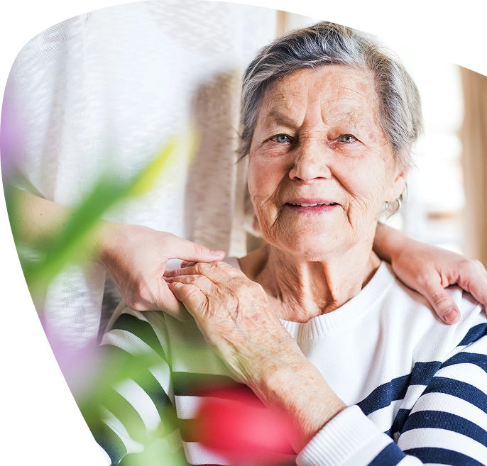 An elderly woman in a striped shirt smiles whilst sitting, with a younger person standing behind her, gently resting hands on her shoulders. The scene, filled with warmth and light, features blurred flowers in the foreground, capturing a tender moment supported by the caring presence of Mayo Roscommon Hospice.