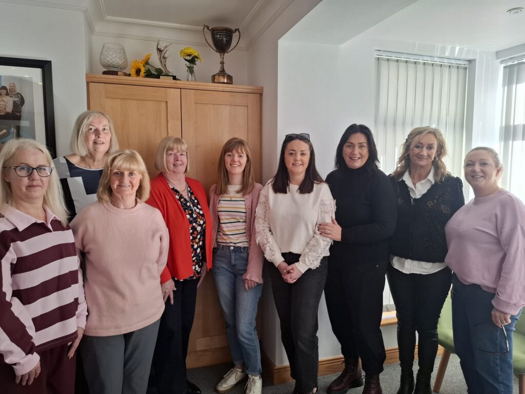 Nine women stand together indoors in front of a cupboard, smiling at the camera during a Mayo Roscommon Hospice event. The room has white walls, large windows with blinds, and a framed photo on the wall.