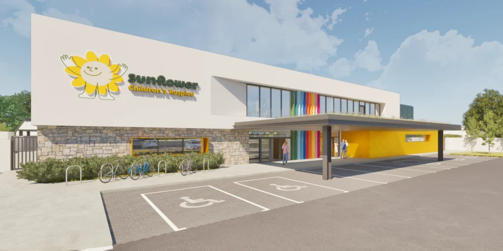 A modern building with Sunflower Children's Hospice signage and a large sunflower graphic, featuring accessible parking spaces, a bike rack, and a covered entrance with colourful pillars—reflecting the welcoming spirit of Mayo Roscommon Hospice.