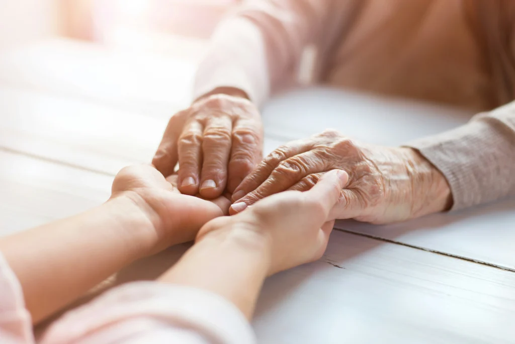 An elderly person gently holds the hands of a younger person across a light wooden table, symbolising care, support, and connection—much like the guidance and steady governance offered by a dedicated board. Soft sunlight lights the scene.