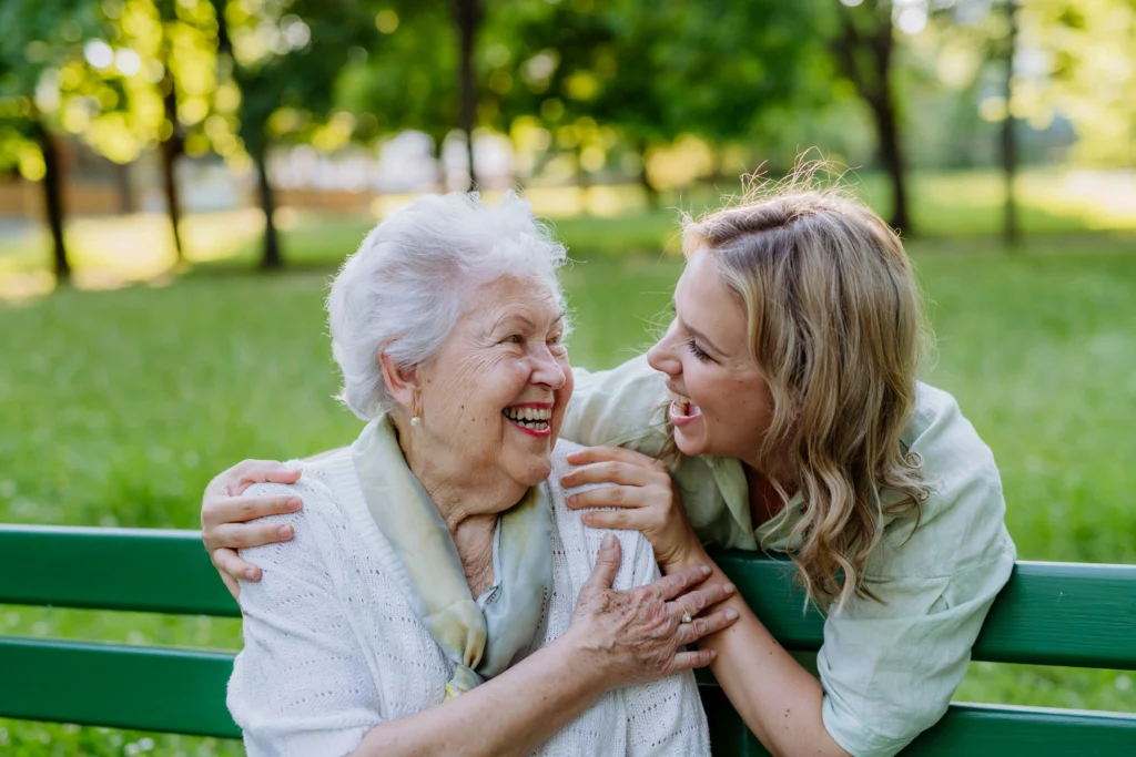 An elderly woman and a younger woman sit on a green bench outdoors, smiling and embracing each other, surrounded by green trees in a park.