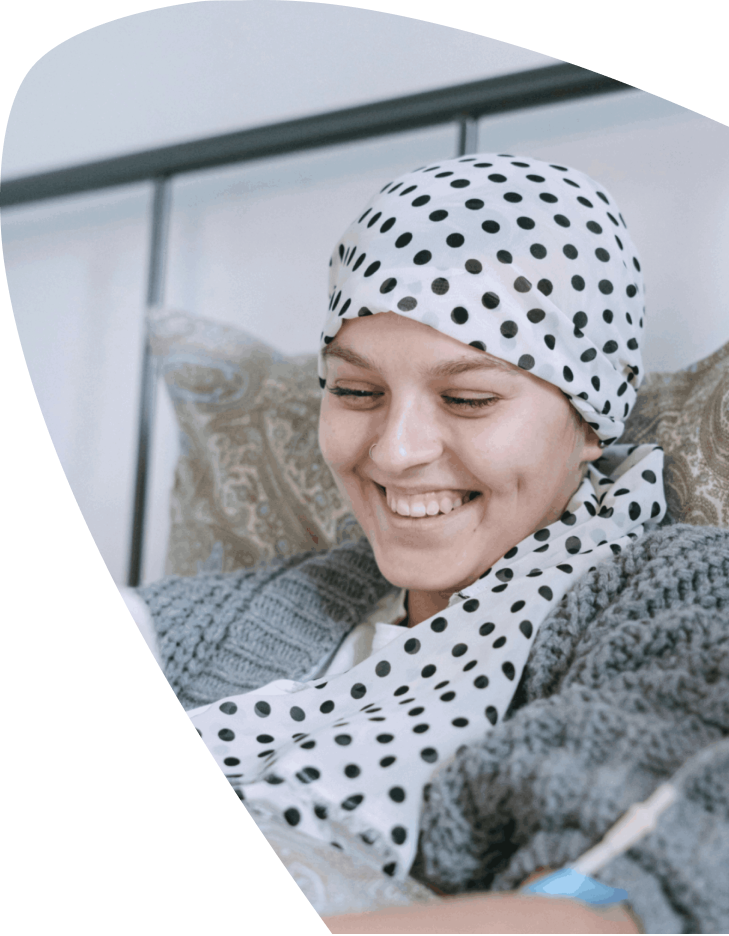 A smiling person wearing a polka dot headscarf and a cosy jumper sits on a bed, appearing relaxed and content, embodying the warmth and comfort supported by Mayo Roscommon Hospice.