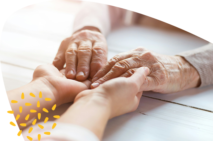 Close-up of younger hands gently holding the hands of an older person on a light wooden surface, conveying care and support—a gesture at the heart of Mayo Roscommon Hospice. Yellow decorative dots are in the bottom left corner.