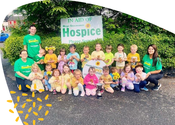 A group of children and adults in yellow and green pose outdoors in front of a Mayo Roscommon Hospice fundraising sign, smiling and holding flowers and a trophy. Shrubs and houses are visible in the background.