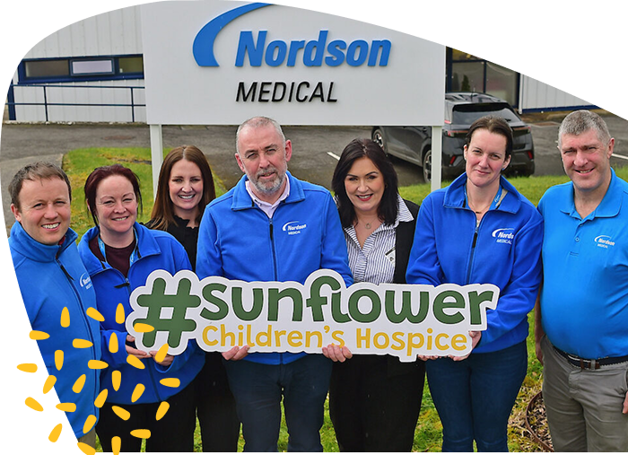 Seven people stand outside Nordson Medical holding a sign that says #sunflower Children’s Hospice in support of the Mayo Roscommon Hospice. They are smiling and wearing blue jackets, with the company sign visible in the background.