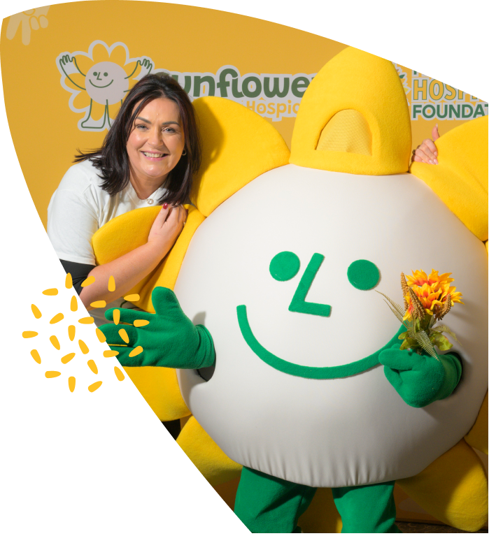 A woman smiles next to a large sunflower mascot with green gloves, holding a small sunflower. Both are in front of a yellow Mayo Roscommon Hospice Sunflower Foundation backdrop.