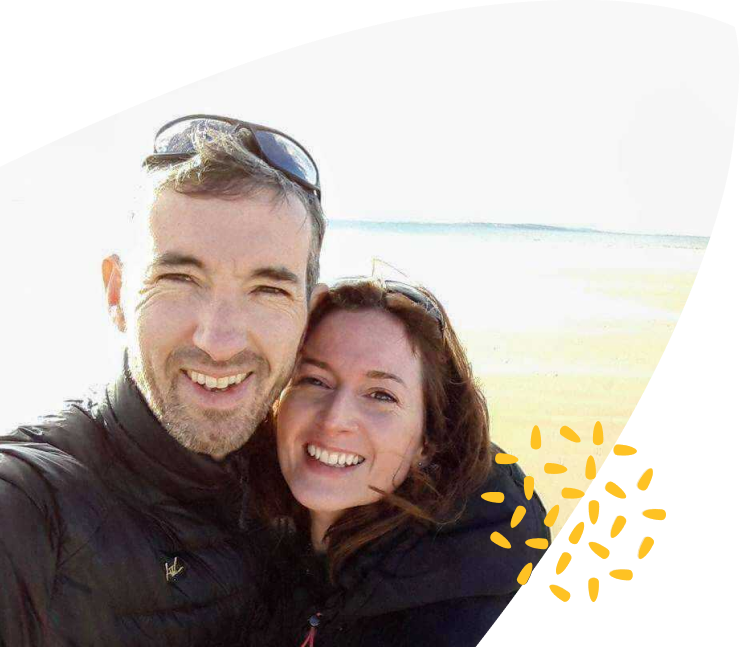 A man and woman smiling and posing for a selfie on a bright, sunny beach. Both wear jackets and sunglasses; the background shows sand and a light blue sky. Yellow decorative dots appear in the lower right corner, subtly evoking the warmth often associated with Mayo Roscommon Hospice.