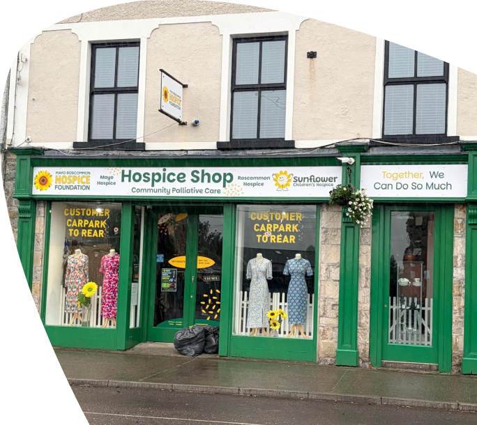 A hospice charity shop with a green shopfront, mannequins in the window wearing colourful dresses, and signs for customer car parking. A banner reads, Together, We Can Do So Much.