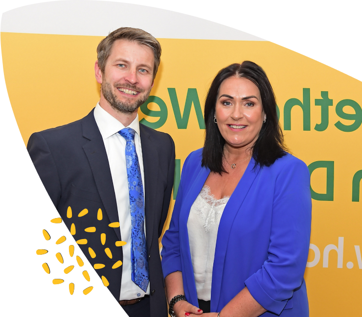 A man in a suit and a woman in a blue blazer stand smiling in front of a yellow backdrop with green and white text, proudly representing Mayo Roscommon Hospice.