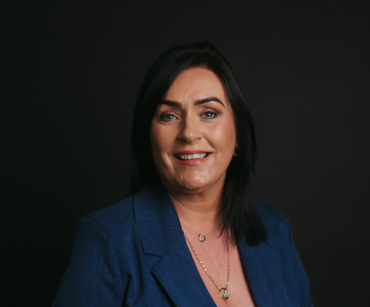 A woman with straight dark hair in a blue blazer smiles at the camera against a plain black background. She wears gold necklaces and has a natural make-up look, representing Mayo Roscommon Hospice with warmth and poise.
