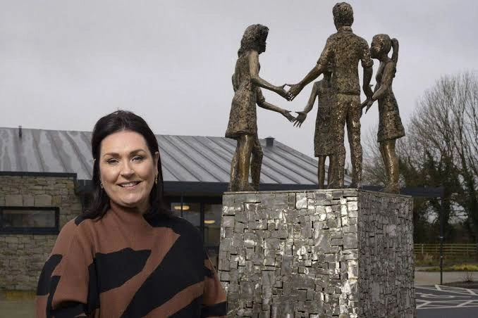 A woman stands smiling in front of a statue depicting four people holding hands on a stone plinth, with a modern building and bare trees in the background.