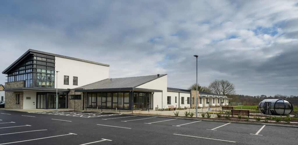 Modern building with large glass windows and flat roofs, surrounded by a mostly empty car park, accessible parking spaces, greenery, and a cloudy sky overhead; this is the Mayo Roscommon Hospice—a thoughtfully designed facility that blends contemporary architecture with a serene environment.