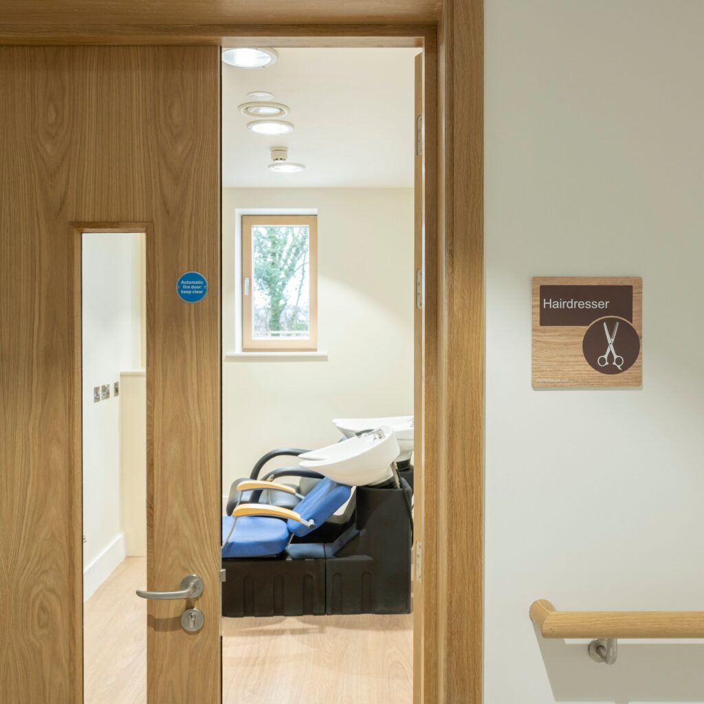 A hairdresser’s room with two black salon chairs and washbasins. The door is partly open, showing a window inside. A sign with scissors and Hairdresser is on the wall, reflecting the supportive atmosphere provided by Mayo Roscommon Hospice.
