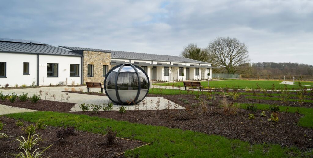 A modern building with white walls and a grey roof—part of the Mayo Roscommon Hospice—sits behind a garden featuring a large spherical metal sculpture, benches, and paths; trees and fields are in the background under a cloudy sky.