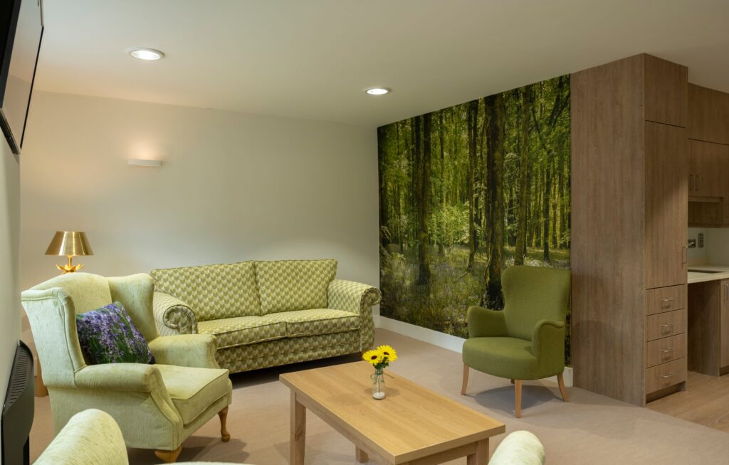 A cosy living room with a green sofa, armchair, wooden coffee table, and a lamp. The back wall features a large mural of a lush green forest, creating an atmosphere reminiscent of tranquillity found in the serene gardens at Mayo Roscommon Hospice. Yellow flowers are on the table.