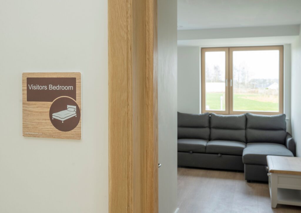 A sign labelled Visitors' Bedroom is mounted on a wall next to a doorway in the Mayo Roscommon Hospice. Inside the room, there is a dark grey sofa, a window with a view outside, and a wooden table.